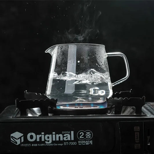Clear glass teapot on a stove with steam rising against a dark background